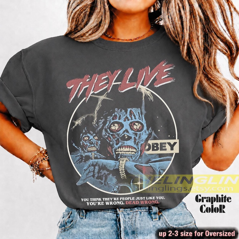 K&ouml;nnte beinhalten: Ein anthrazitfarbenes Kurzarm-T-Shirt mit einem Grafikdruck von zwei skelettierten Figuren in einem Kreis. Der Text lautet "THEY LIVE" oben, "OBEY" rechts und "YOU THINK THEY'RE PEOPLE JUST LIKE YOU. YOU'RE WRONG. DEAD WRONG." am unteren Rand.