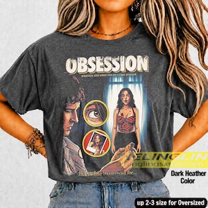 K&ouml;nnte beinhalten: Dunkelgraues meliertes T-Shirt mit dem Wort "OBSESSION" im Vintage-Stil. Das Shirt zeigt ein Filmplakat-Design mit Illustrationen eines Mannes, einer Frau und einem Polaroid-Foto. Der Text "Be careful who you wish for..." ist ebenfalls sichtbar.