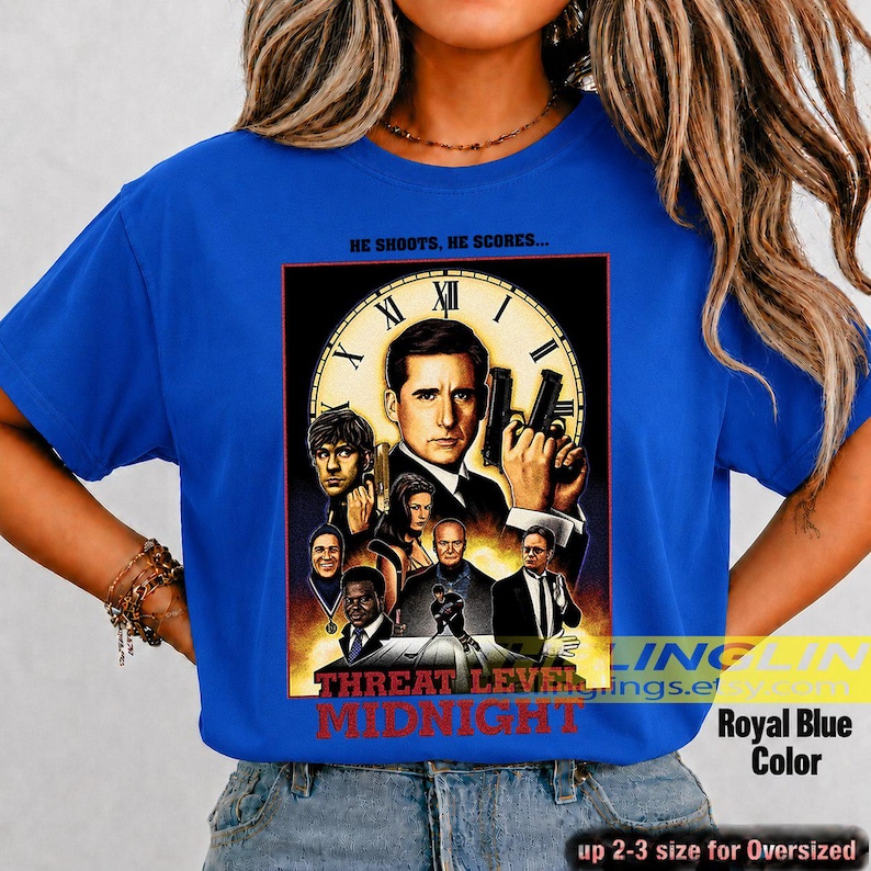 May include: A royal blue short-sleeve t-shirt featuring a graphic print of a movie poster titled Threat Level Midnight. The design includes a large clock face, multiple characters, and the text He Shoots, He Scores at the top and bottom.