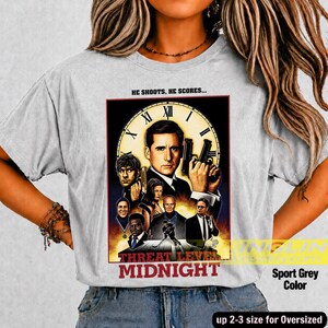 May include: A light gray t-shirt featuring a graphic print of a clock face background with multiple character portraits and the text "HE SHOOTS, HE SCORES..." at the top and "MIDNIGHT" at the bottom, arranged in a movie poster style layout.