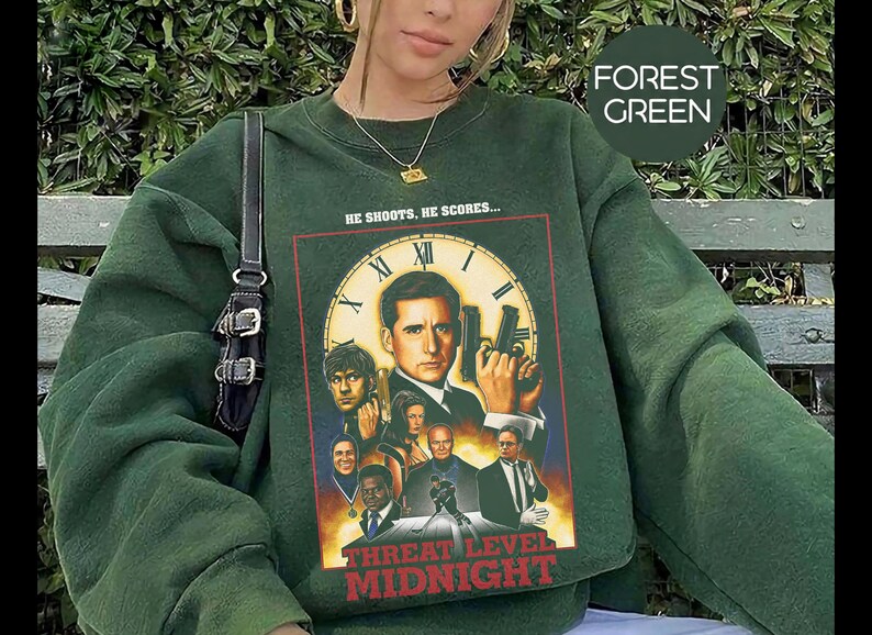 May include: A forest green crewneck sweatshirt featuring a graphic print of the movie poster for Threat Level Midnight. The design includes a central clock face, multiple characters, and the text HE SHOOTS, HE SCORES... and THREAT LEVEL MIDNIGHT.