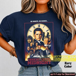 May include: A navy blue t-shirt featuring a graphic print of the movie poster for Threat Level Midnight. The design includes a large clock face, multiple character portraits, and the text HE SHOOTS, HE SCORES... and THREAT LEVEL MIDNIGHT.