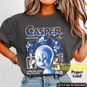 May include: Dark gray t-shirt featuring the movie title "Casper" in white and blue lettering, with a graphic of Casper the friendly ghost and other characters. The shirt also includes the text "Seeing is Believing..." and the names "Christina Ricci" and "Bill Pullman".