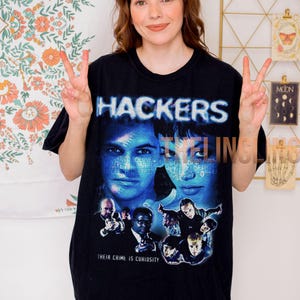 Puede incluir: Camiseta negra con el título de la película "Hackers" en azul y blanco, y un gráfico del elenco. La frase "Their Crime Is Curiosity" está impresa debajo. La camiseta la lleva una persona.