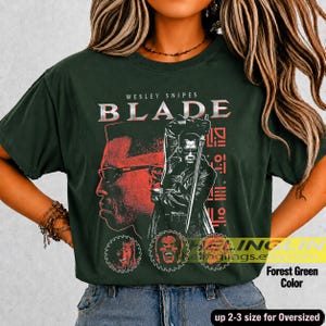 May include: A forest green short-sleeve t-shirt featuring a graphic print of Wesley Snipes as Blade. The design includes the text BLADE, a large profile silhouette, a central figure holding a sword, and three circular character portraits.