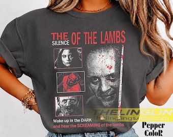 May include: A charcoal grey t-shirt featuring a graphic print from The Silence of the Lambs. The design includes three small framed images and a large portrait of a face with red splatter, accompanied by the text The Silence of the Lambs.