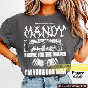May include: A charcoal grey short-sleeve t-shirt featuring a white graphic print. The design includes the text "MANDY" above a close-up of eyes, the phrase "I COME FOR THE REAPER," a small cross, and the text "I'M YOUR GOD NOW" at the bottom.