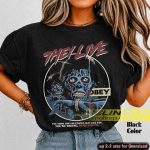 K&ouml;nnte beinhalten: Ein schwarzes Kurzarm-T-Shirt mit einem Grafikdruck aus dem Film They Live. Das Design zeigt eine blau get&ouml;nte Alien-Figur in einem wei&szlig;en Kreis mit dem Text They Live, Obey und You think they're people just like you. You're wrong. Dead wrong.