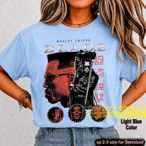 May include: A light blue t-shirt featuring a graphic print of Wesley Snipes as Blade. The design includes the text BLADE, a large profile of the character, a figure holding a sword, and three circular portraits at the bottom in red and black.