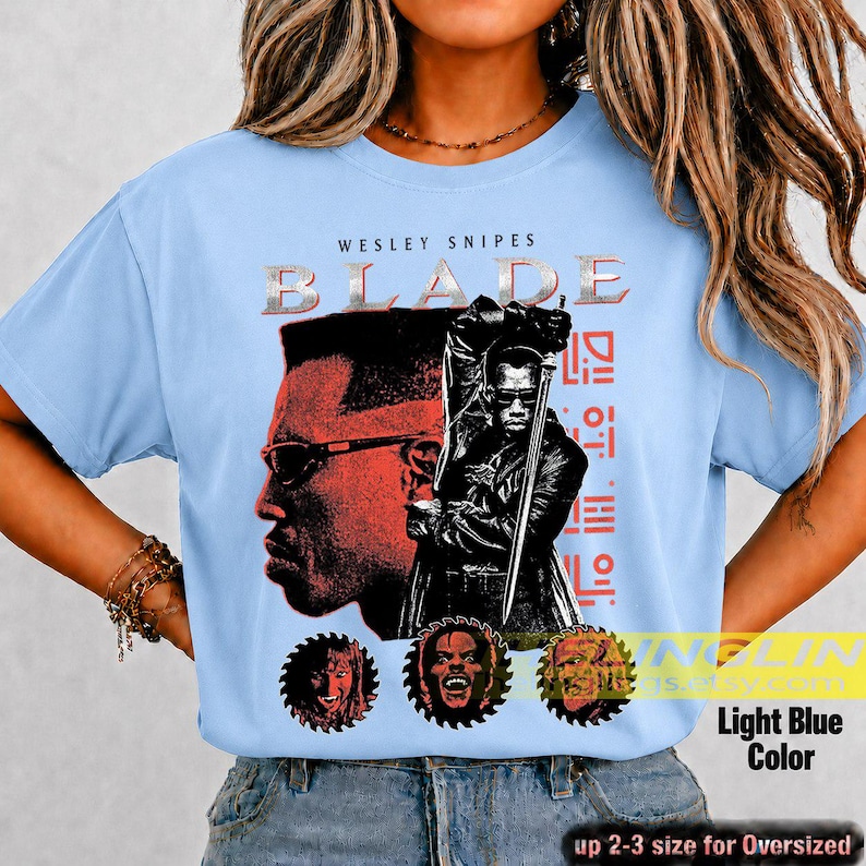 May include: A light blue t-shirt featuring a graphic print of Wesley Snipes as Blade. The design includes the text BLADE, a large profile of the character, a figure holding a sword, and three circular portraits at the bottom in red and black.