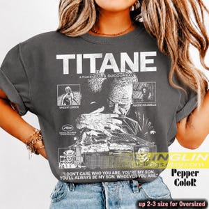 May include: A charcoal gray short-sleeve t-shirt featuring a white graphic print inspired by the film Titane. The design includes the title, cast names, and the quote: I DON'T CARE WHO YOU ARE. YOU'RE MY SON. YOU'RE ALWAYS MY SON.