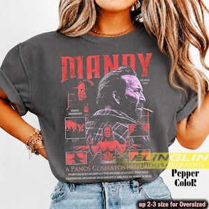 May include: Dark gray t-shirt featuring the movie title "MANDY" in red, along with images of actors and the text "A PANOS COSMATOS PICTURE". The shirt is labeled "Pepper ColoR" and is available in up to 2-3 sizes for an oversized fit.