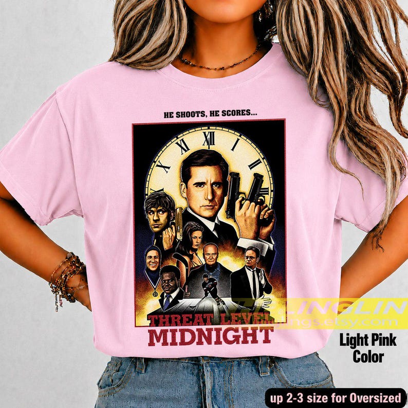 May include: A light pink t-shirt featuring a graphic print of a movie-style poster with the text HE SHOOTS, HE SCORES... and THREAT LEVEL MIDNIGHT. The design includes a large clock face background and multiple character portraits in a collage.