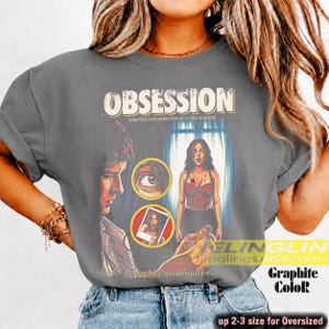 K&ouml;nnte beinhalten: Graphitgraues &uuml;bergro&szlig;es T-Shirt mit einem Filmplakat-Design. Das Design zeigt das Wort "OBSESSION" in einer Vintage-Schriftart, zusammen mit Illustrationen eines Mannes, einer Frau und einer Nahaufnahme eines Auges. Der Text "Be careful who you wish for..." ist ebenfalls enthalten.
