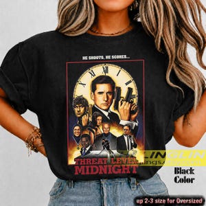 May include: A black t-shirt featuring a graphic print of a movie poster titled Threat Level Midnight. The design includes a large clock face, multiple character portraits, and the text HE SHOOTS, HE SCORES... above the central figure holding a gun.