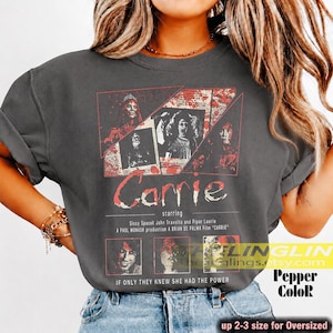 Può includere: Una t-shirt grigio antracite con una stampa grafica ispirata al film Carrie. Il design include testo rosso e bianco, diverse immagini di scene del film e i nomi degli attori Sissy Spacek, John Travolta e Piper Laurie stampati sul davanti.