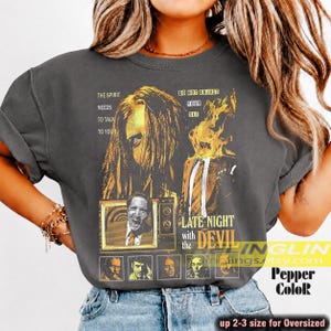 May include: A charcoal gray t-shirt featuring a graphic print with the text "THE SPIRIT NEEDS TO TALK TO YOU", "DO NOT ADJUST YOUR SET", and "LATE NIGHT with the DEVIL". The design includes a stylized portrait, a television screen, and flames.