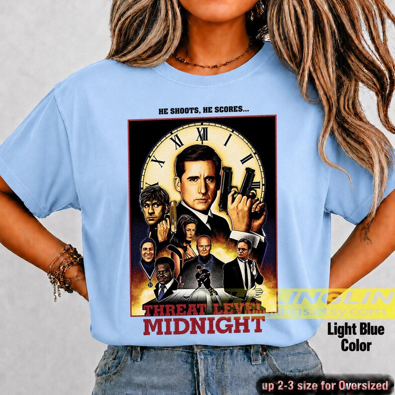 May include: A light blue t-shirt featuring a graphic print of the movie poster Threat Level Midnight. The design includes a large clock face, multiple character portraits, and the text HE SHOOTS, HE SCORES... and THREAT LEVEL MIDNIGHT.