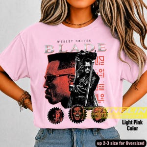 May include: A light pink t-shirt featuring a graphic print of Wesley Snipes as Blade. The design includes the text 'WESLEY SNIPES BLADE', a large portrait, a figure holding a sword, and three circular character icons at the bottom.