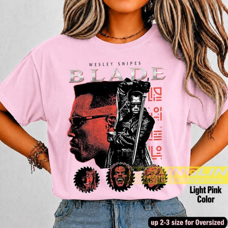 May include: A light pink t-shirt featuring a graphic print of Wesley Snipes as Blade. The design includes the text 'WESLEY SNIPES BLADE', a large portrait, a figure holding a sword, and three circular character icons at the bottom.