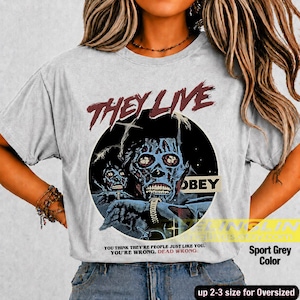 K&ouml;nnte beinhalten: Ein hellgraues T-Shirt mit einem Grafikdruck einer skelettartigen Figur mit dunkelblauer Haut und einer Armbanduhr. Der Text lautet THEY LIVE in Rot, OBEY in einem schwarzen Kasten und YOU THINK YOU ARE PEOPLE JUST LIKE YOU. YOU ARE WRONG. DEAD WRONG.