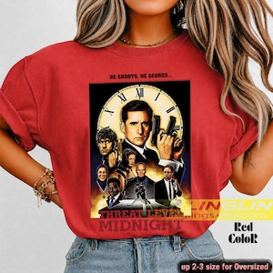 May include: A red t-shirt featuring a graphic print of the movie poster for Threat Level Midnight. The design includes a large clock face background, a central male figure holding a gun, and several smaller character portraits below.