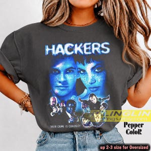 Peut inclure: T-shirt gris foncé avec le mot "HACKERS" en bleu, et un graphisme bleuté de deux visages et d'autres personnages. Le texte "THEIR CRIME IS CURIOSITY" est en dessous. Le t-shirt est surdimensionné.