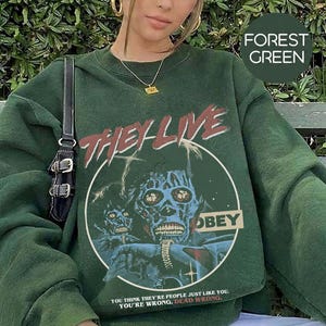 K&ouml;nnte beinhalten: Ein waldgr&uuml;nes Sweatshirt mit Rundhalsausschnitt und einem Grafikdruck mit dem Text THEY LIVE und OBEY. Das Design enth&auml;lt eine bl&auml;uliche Illustration einer totenkopfartigen Figur und den Text YOU THINK THEY'RE PEOPLE JUST LIKE YOU. YOU'RE WRONG. DEAD WRONG.