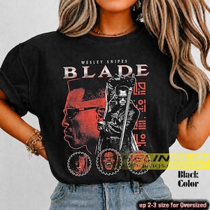 May include: A black short-sleeve t-shirt featuring a graphic print of Wesley Snipes as Blade. The design includes the text BLADE, a large profile portrait, a figure holding a sword, and three circular character icons in red and white.