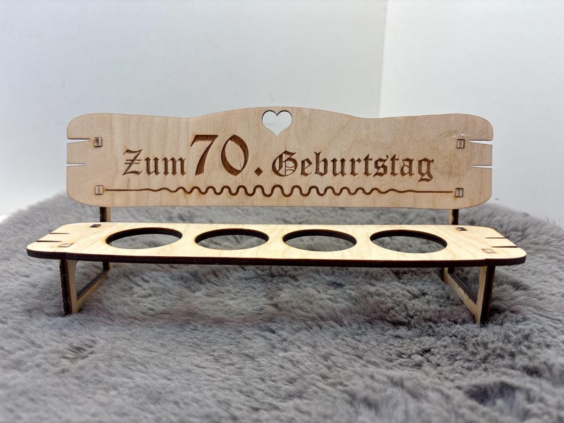 May include: Wooden bench with four slots for holding eggs. The bench has a heart-shaped cutout and the text "Zum 70. Geburtstag" is engraved on the back.