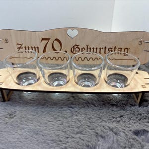 May include: Wooden shot glass holder with the text "Zum 70. Geburtstag" and a heart. The holder has four shot glasses.