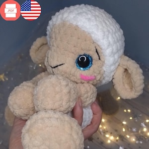 May include: A crocheted white and beige plush lamb toy with blue glitter eyes and a pink blush on its cheek. The lamb is sitting with its head tilted to the side and one eye closed.