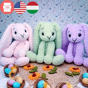 May include: Three crocheted bunny rabbit plush toys in pink, green, and purple. The bunnies are sitting on a woven straw mat with small yellow felt flowers and nests with colorful eggs.