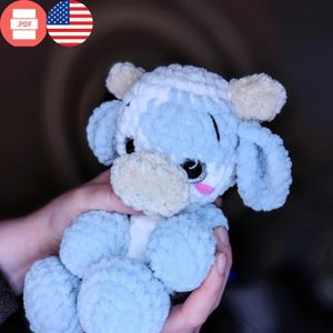 Puede incluir: Un juguete de peluche de vaca azul y blanco tejido a crochet con una nariz rosa y ojos brillantes. El juguete está siendo sostenido por la mano de una persona.