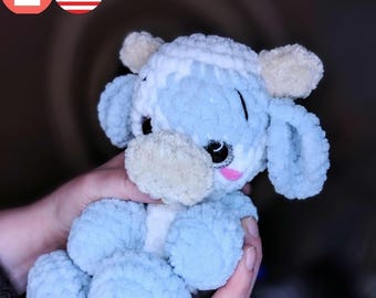 May include: A crocheted blue and white cow plush toy with a pink nose and sparkly eyes. The toy is being held by a person's hand.