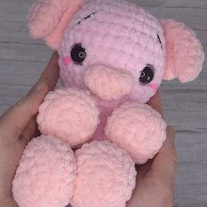 Puede incluir: Un pequeño cerdo de peluche rosa tejido a crochet con ojos negros y un pequeño hocico rosa.