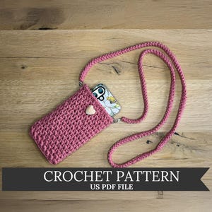 May include: A pink crocheted phone case with a matching strap, featuring a wooden heart-shaped button closure. The phone case holds a phone with a floral design. The text "CROCHET PATTERN" is displayed on a banner below the case.