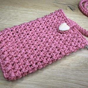 May include: A handmade, pink crochet phone bag with a matching strap. The bag features a wooden heart-shaped button closure and a silver-tone clasp. The bag is made of a thick, textured yarn.