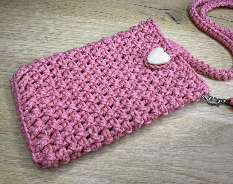 May include: A handmade, pink crochet phone bag with a matching strap. The bag features a wooden heart-shaped button closure and a silver-tone clasp. The bag is made of a thick, textured yarn.