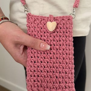 May include: A dusty rose crochet phone bag with a matching strap and a wooden heart accent. The bag is held by a person. The bag has a woven texture and silver-colored metal clasps.