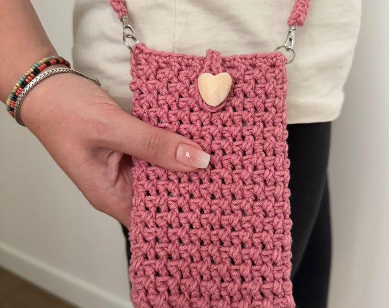 May include: A dusty rose crochet phone bag with a matching strap and a wooden heart accent. The bag is held by a person. The bag has a woven texture and silver-colored metal clasps.