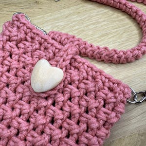 May include: A handmade, pink crocheted crossbody bag with a matching strap and a wooden heart accent. The bag features silver-tone metal clasps and a thick, textured stitch. The bag is designed for carrying small items.