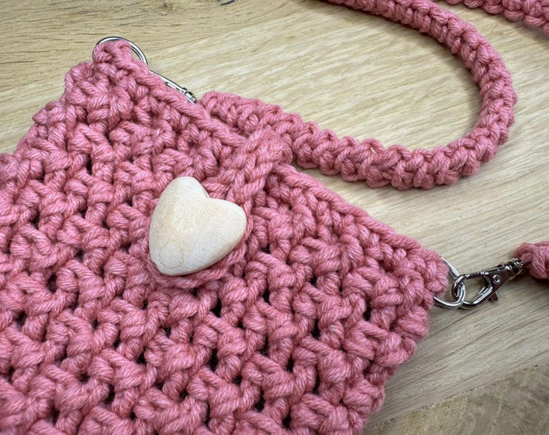May include: A handmade, pink crocheted crossbody bag with a matching strap and a wooden heart accent. The bag features silver-tone metal clasps and a thick, textured stitch. The bag is designed for carrying small items.