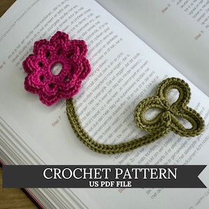 Pode incluir: Um marcador de livro de crochê com uma flor rosa e um trevo verde. O marcador está sobre um livro com o texto "CROCHET PATTERN US PDF FILE" impresso nele.