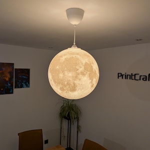May include: A spherical pendant light fixture featuring a detailed, textured surface that mimics the appearance of the moon. The lamp is suspended from the ceiling by a white cord and a white canopy, emitting a warm, soft glow from within.