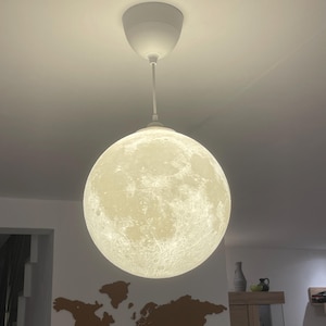 Pode incluir: Uma luminária pendente esférica com uma superfície detalhada e texturizada que imita a aparência da lua. A lâmpada está suspensa no teto por um fio branco e um suporte circular branco correspondente, emitindo um brilho suave.