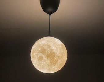 May include: A spherical pendant light fixture featuring a textured, glowing surface that mimics the appearance of the moon. The lamp is suspended by a thin black cord from a matching black circular ceiling canopy, emitting a warm light.