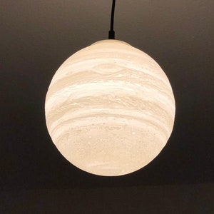 Eine kugelförmige Pendelleuchte mit einer strukturierten, cremeweißen Oberfläche und horizontalen Bändern Sie stellt den Jupiter dar, die an das Aussehen eines Planeten erinnern. Die Kugel hängt an einem dünnen schwarzen Kabel an einem schwarzen, runden Deckenbaldachin.
