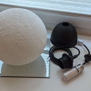 Eine weiße, kugelförmige Mondlampe mit texturierter, kraterartiger Oberfläche, die auf einem quadratischen Spiegel ruht. Daneben befinden sich eine schwarze Pendelleuchtenfassung, ein schwarzes Stromkabel und eine zylindrische LED-Lampe.