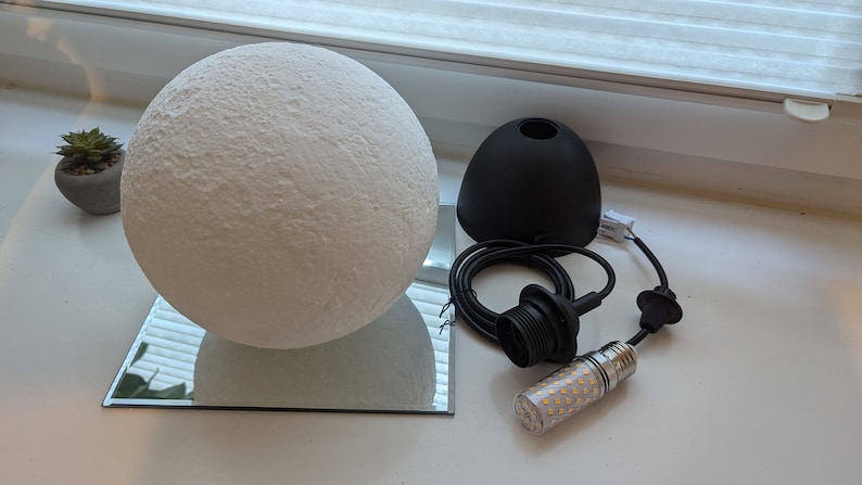 Eine weiße, kugelförmige Mondlampe mit texturierter, kraterartiger Oberfläche, die auf einem quadratischen Spiegel ruht. Daneben befinden sich eine schwarze Pendelleuchtenfassung, ein schwarzes Stromkabel und eine zylindrische LED-Lampe.